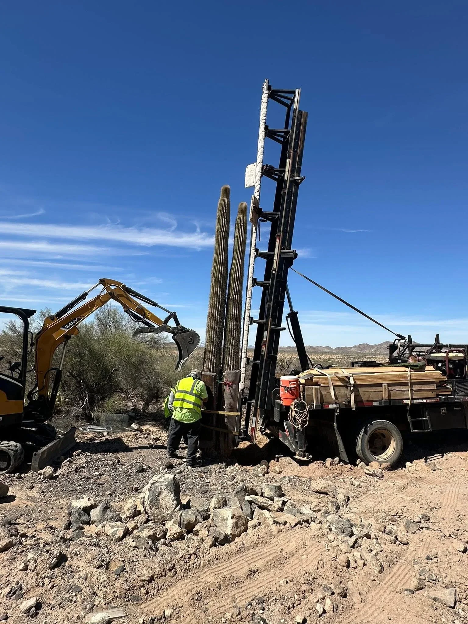 Hydraulic lift truck removing large saguaro cactus with excavator and crew on rocky desert site